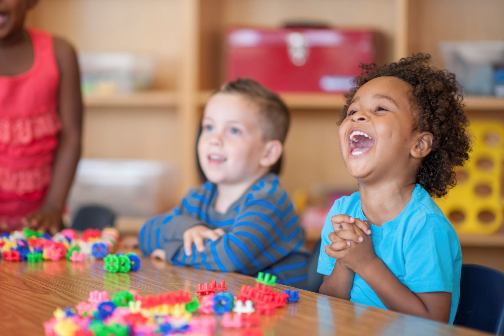 Preschool children are playing with toy blocks and laughing together at a classroom table.
